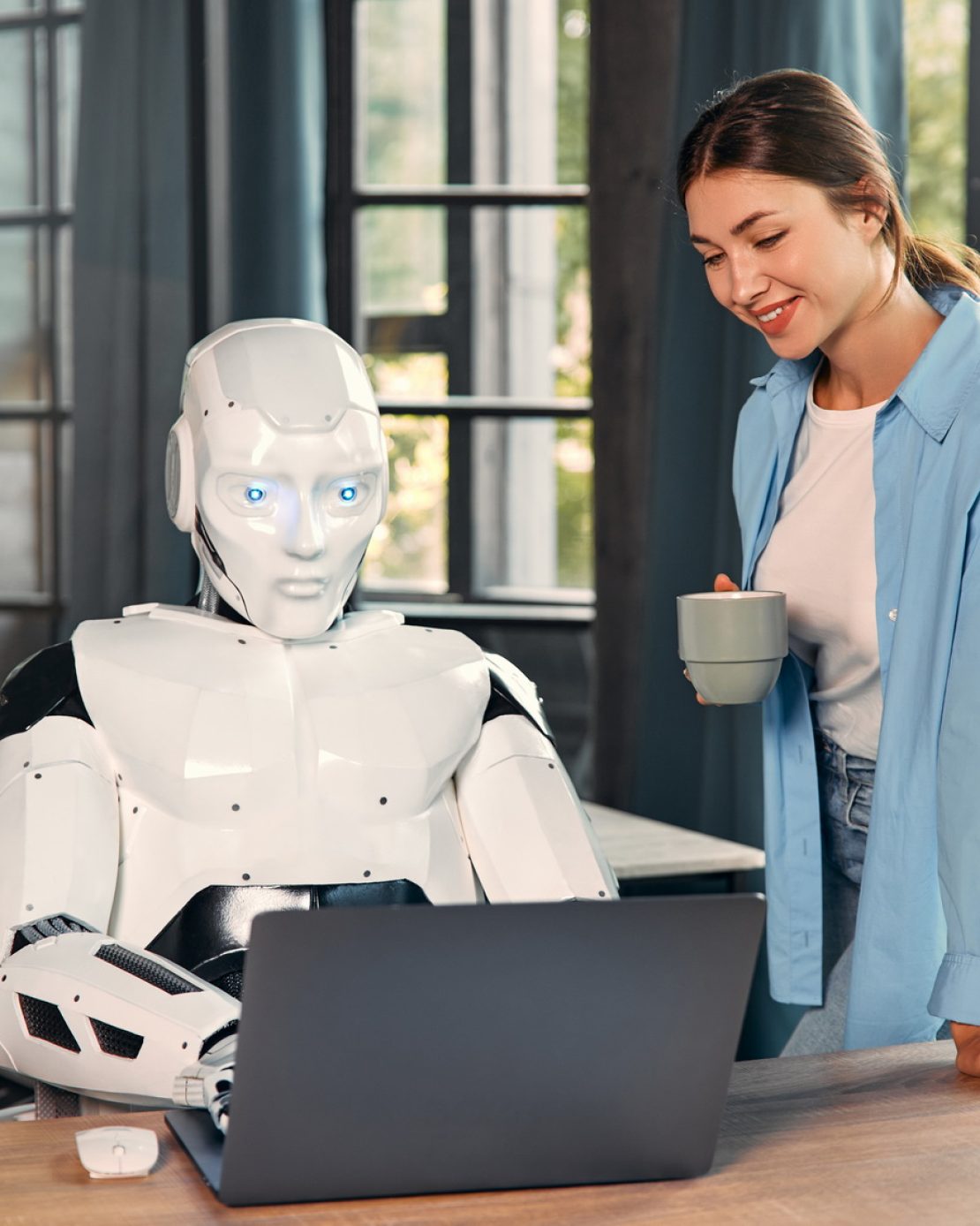 Young woman with a cup of coffee and a humanoid robot working while sitting at a laptop in a modern office. Collaboration between humans and artificial intelligence.